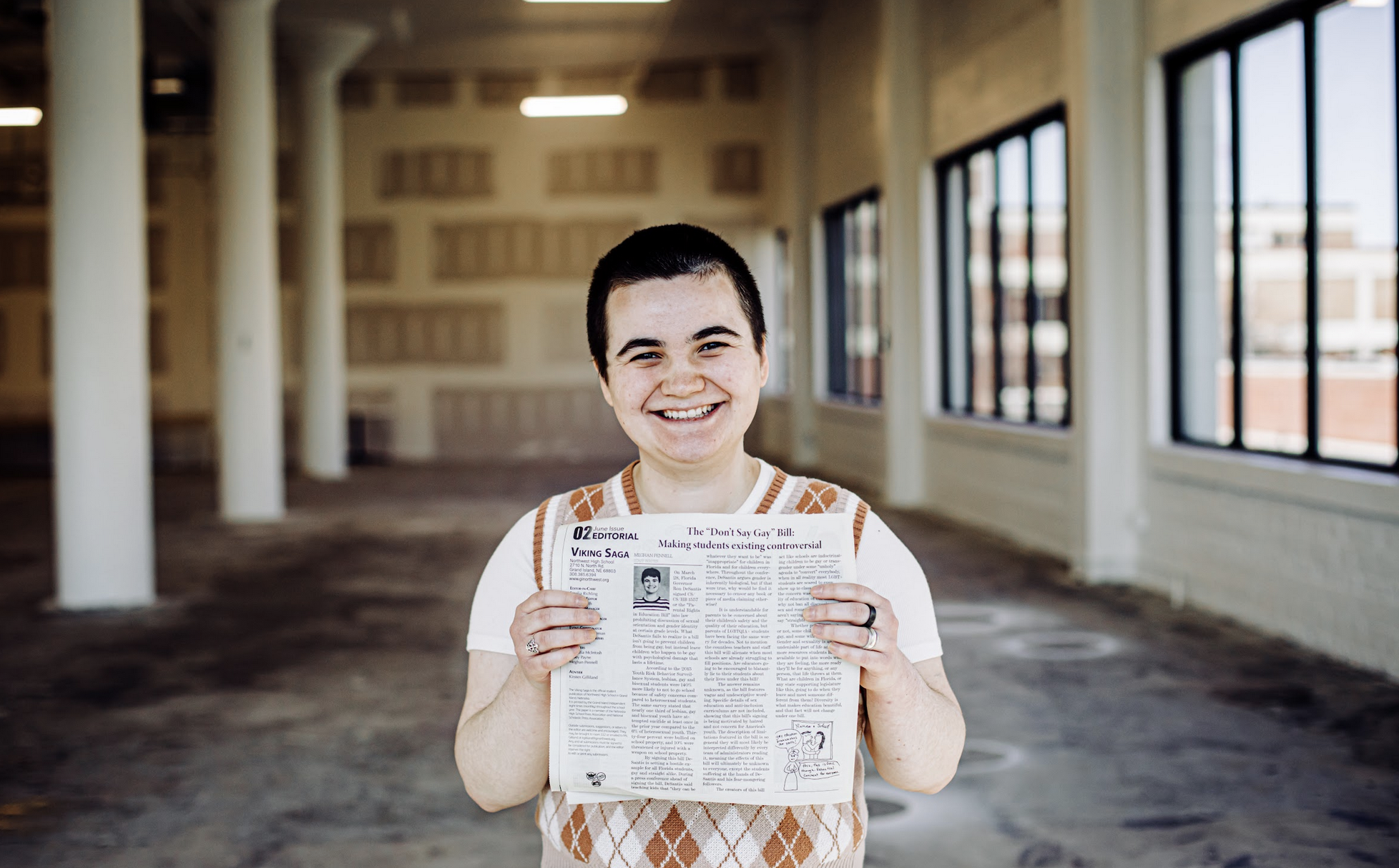 A photo of a smiling student who holds a newspaper in front of themself. 